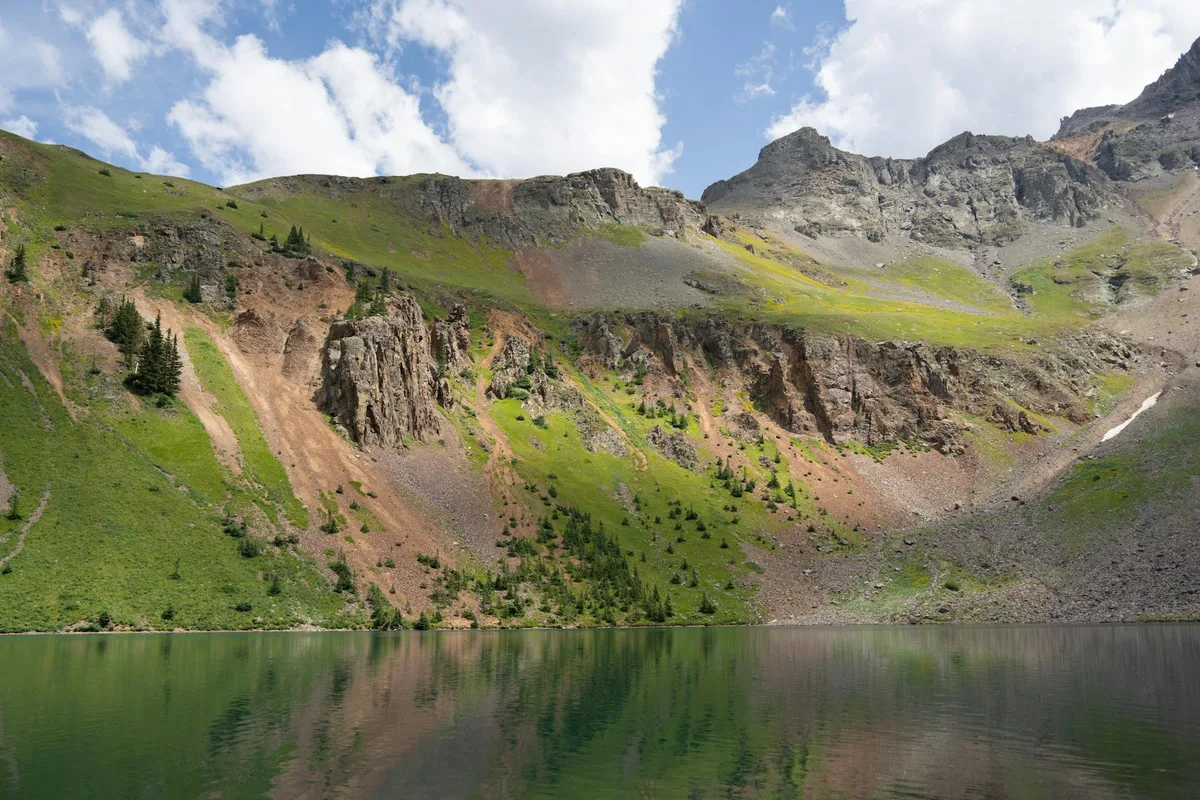 Calm lake water fills the foreground, mirroring steep gray cliffs, green alpine slopes, and scattered conifers under a bright blue sky with puffy clouds. The rugged mountain backdrop and patchy summer greenery create a serene, wide landscape reflection.