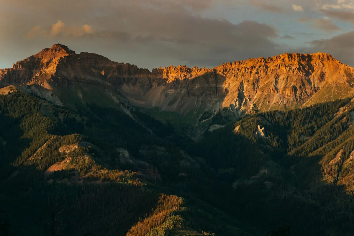 Golden hour light washes over rugged mountain peaks and steep forested slopes, with warm orange highlights along the ridgeline and deep shadows filling the valleys below. The scene feels expansive and dramatic, with layered terrain fading into the distance beneath a partly cloudy sky.