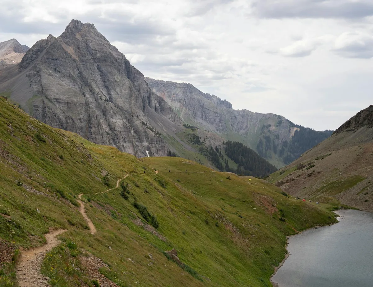 A narrow dirt trail winds through green alpine slopes beside a still mountain lake, with jagged gray peaks rising sharply in the background under a cloudy sky. Small tents dot the hillside, adding scale to the broad, rugged landscape.