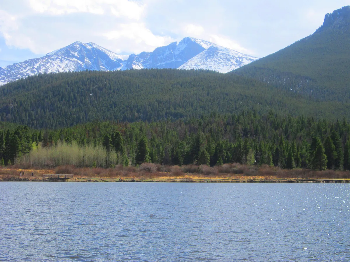 Snow-dusted Longs Peak and Mount Meeker rise above a dark evergreen ridge, reflected in the calm blue surface of Lily Lake. A narrow strip of shoreline with early spring shrubs and a small viewing deck runs along the far side under bright, hazy light.