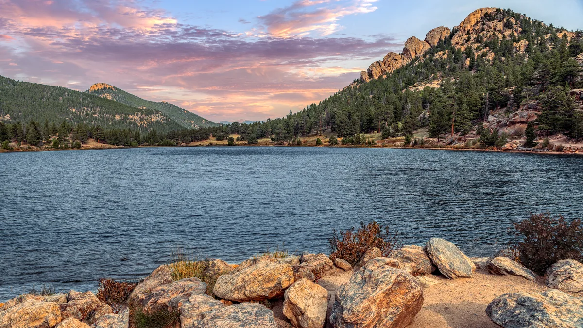 Rocky shoreline boulders frame Lily Lake in the foreground, with dark water stretching toward pine-covered hills and sunlit granite cliffs under a pink-and-gold sunset sky. The scene feels calm and expansive, with warm light catching the mountain ridge and faint ripples across the lake.