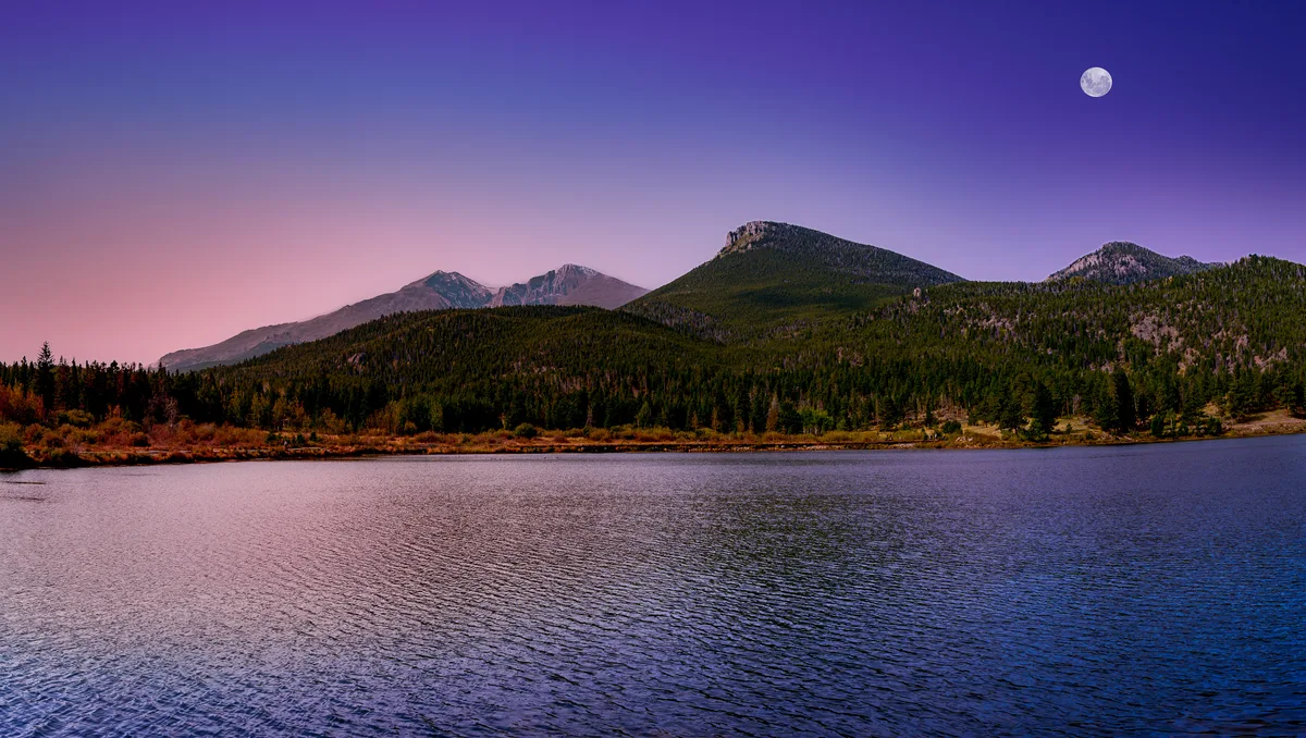 A twilight view across Lily Lake shows rippled water in the foreground, a dark evergreen shoreline, and the jagged silhouette of Longs Peak and nearby ridges under a pink-to-deep-blue sky. The full moon hangs above the mountains, adding a calm, dramatic glow to the scene.