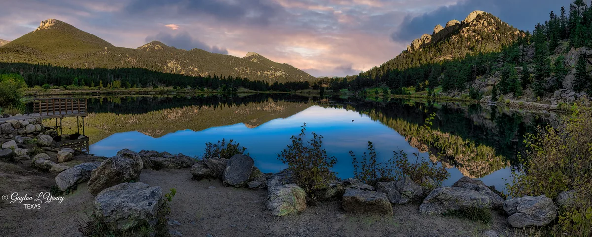 Golden-hour light warms the rocky mountain slopes above Lily Lake, with Longs Peak and nearby peaks mirrored in the still water. A stone-lined shoreline and small viewing platform frame the foreground, while the lake’s glassy surface creates a near-symmetrical reflection.