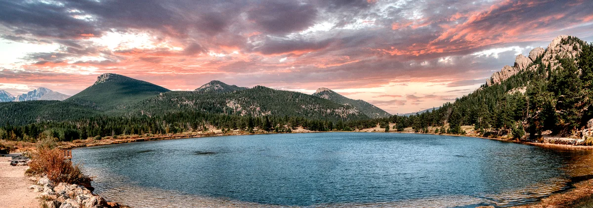 Wide view of Lily Lake at golden hour, with calm blue water in the foreground, rocky shoreline and pines along the edges, and Longs Peak and nearby granite ridges rising under a pink-and-orange sky. The lake’s surface is mostly clear, with soft ripples reflecting the warm sunset light.