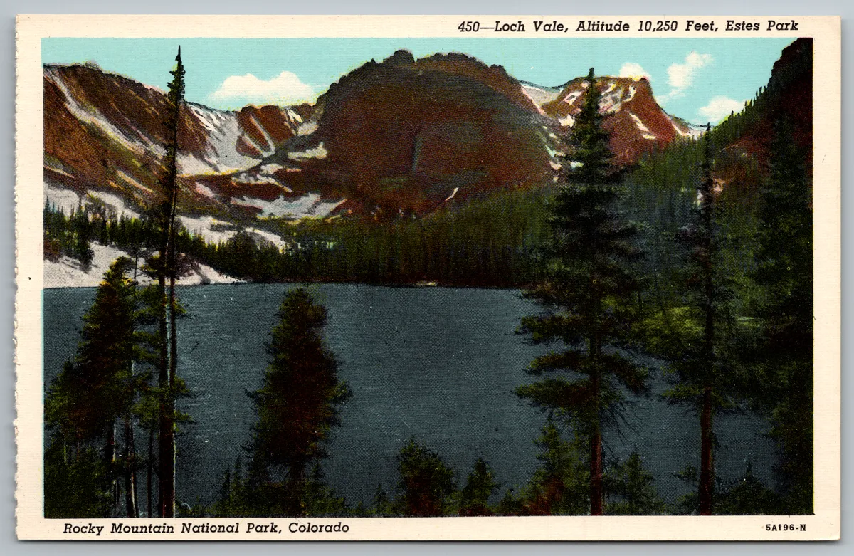 Emerald alpine water fills the center of the scene, framed by dark evergreen trees in the foreground and steep granite peaks with lingering snow under a clear morning sky. The lake sits in a broad U-shaped glacial valley, with bright white snow patches and rugged cliffs rising above the far shore.