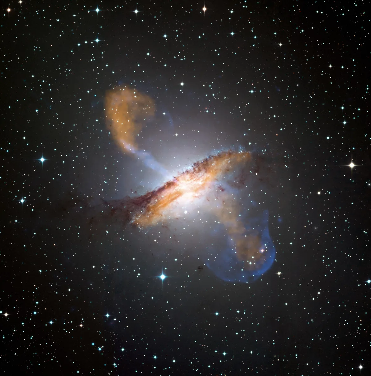 A deep-space scene filled with countless stars surrounds a bright, glowing galaxy at center, with orange and blue plumes arcing outward like jet outflows. The galaxy’s dusty, luminous core stands out sharply against the black background.