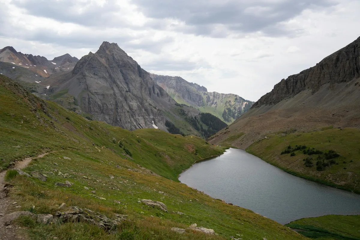 A narrow alpine lake curves through a steep glacial valley, surrounded by green tundra slopes and towering gray granite cliffs. Cloudy morning light softens the scene, with patches of lingering snow visible on the distant peaks.