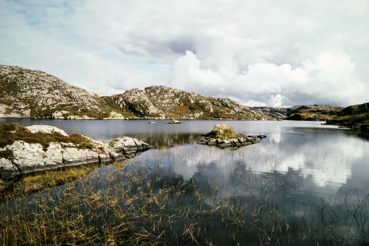 Calm alpine lake water reflects bright morning clouds above low, rugged granite hills. Marsh grasses and pale boulders frame the foreground, with a few small rocky islets breaking the glassy surface.