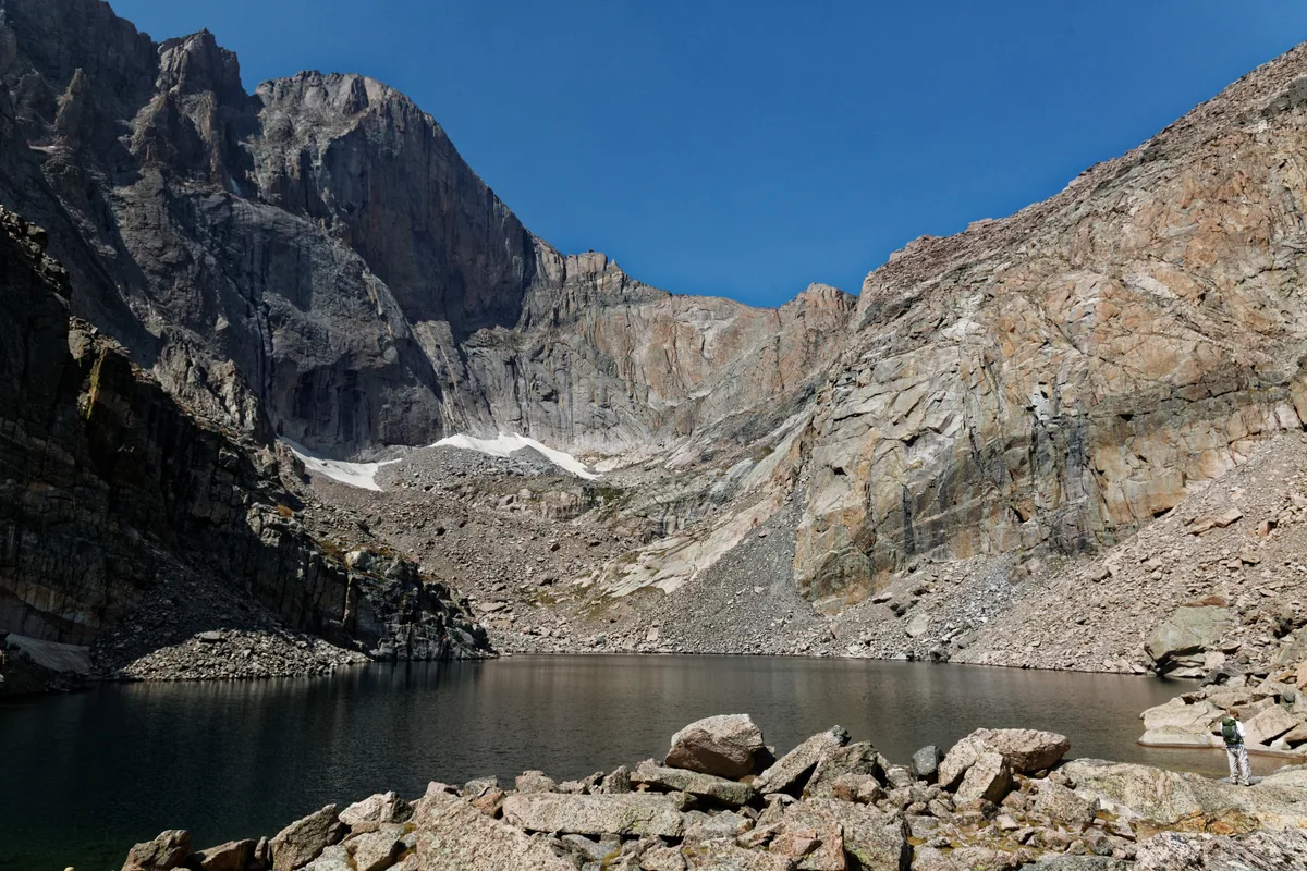 Chasm Lake sits in a rocky alpine cirque beneath the sheer granite walls of Longs Peak’s Diamond, with calm dark water reflecting the cliffs. Sunlit talus slopes and a small patch of lingering snow contrast with the deep blue morning sky.
