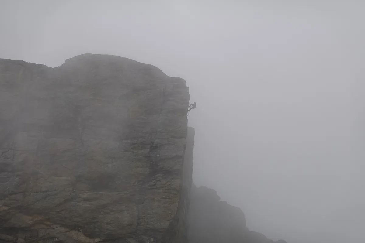 A sheer dark granite wall rises out of thick morning fog, with a tiny climber silhouetted near the narrow edge of the cliff. The rest of the mountain fades into mist, emphasizing the scale and exposure of the alpine face.