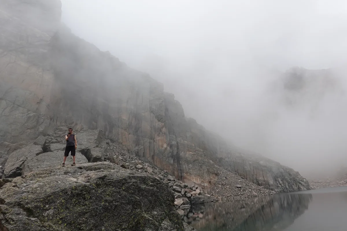 Mist hangs over Chasm Lake beneath the towering gray cliff of Longs Peak’s Diamond, with the calm water reflecting the rock face. A lone hiker stands on a boulder in the foreground, emphasizing the scale of the steep alpine cirque.