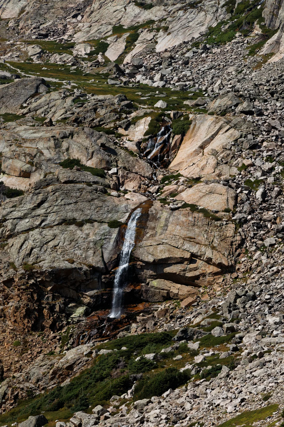 A narrow waterfall spills down a steep granite chute into a rocky alpine basin, with pale, striated rock slabs and scattered patches of green tundra. The vertical composition emphasizes the rugged, high-elevation landscape under bright morning light.