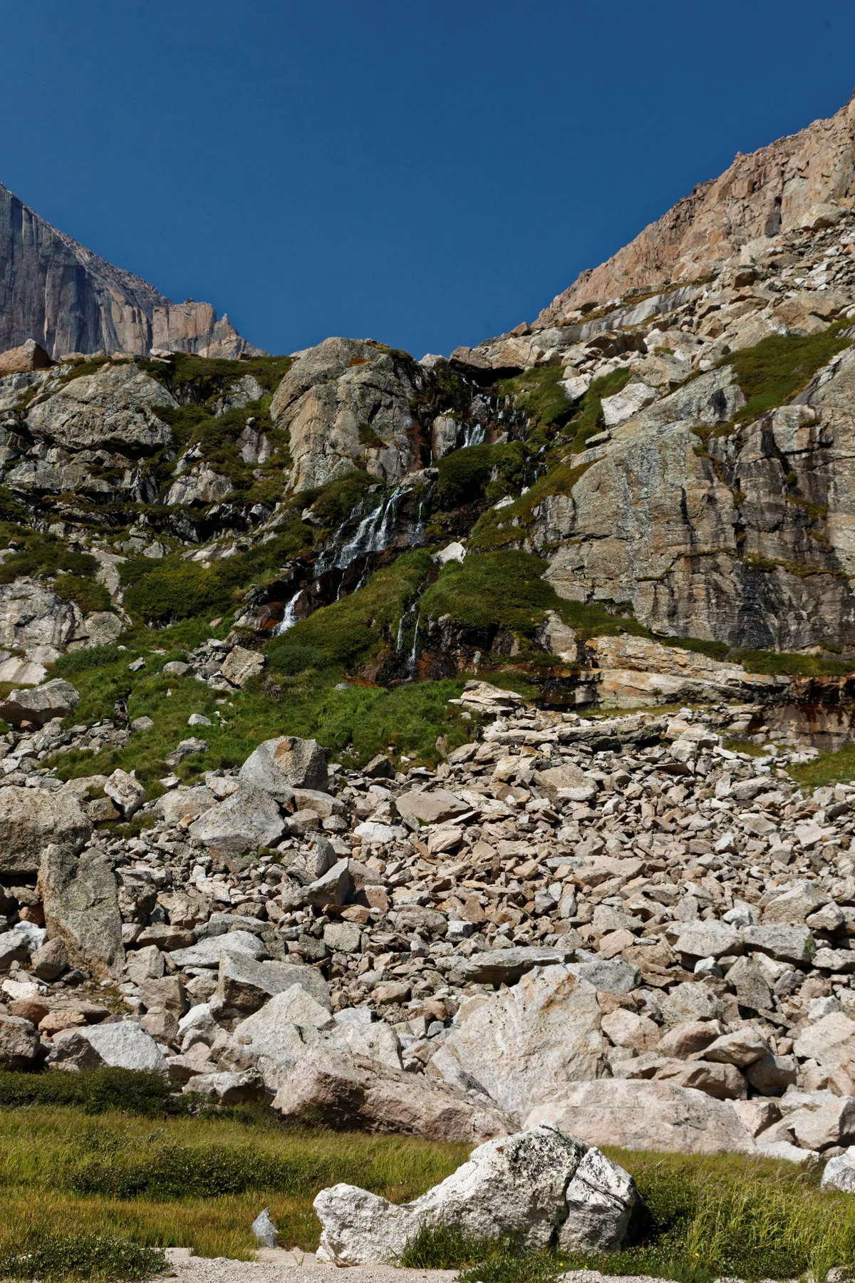 Steep granite cliffs rise above a rocky alpine slope dotted with patches of green tundra, with a small waterfall spilling down the center under a clear deep-blue sky. The left edge of the Diamond face of Longs Peak peeks into view, emphasizing the high, rugged cirque.