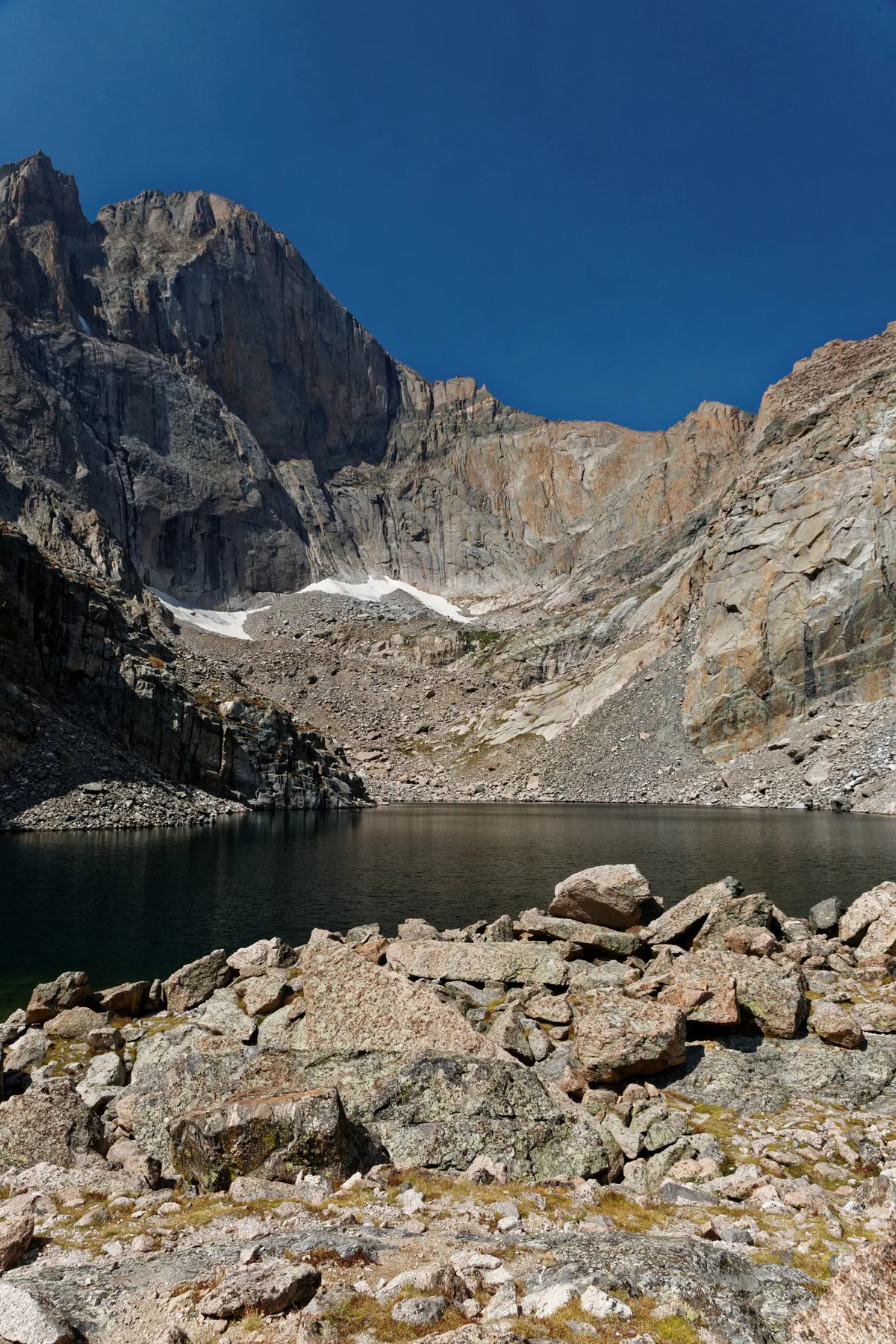 Chasm Lake lies still below the sheer gray cliffs of the Diamond, with Longs Peak’s massive east face rising under a clear deep-blue sky. Rocky talus and a few lingering snow patches frame the dark alpine water in the foreground.