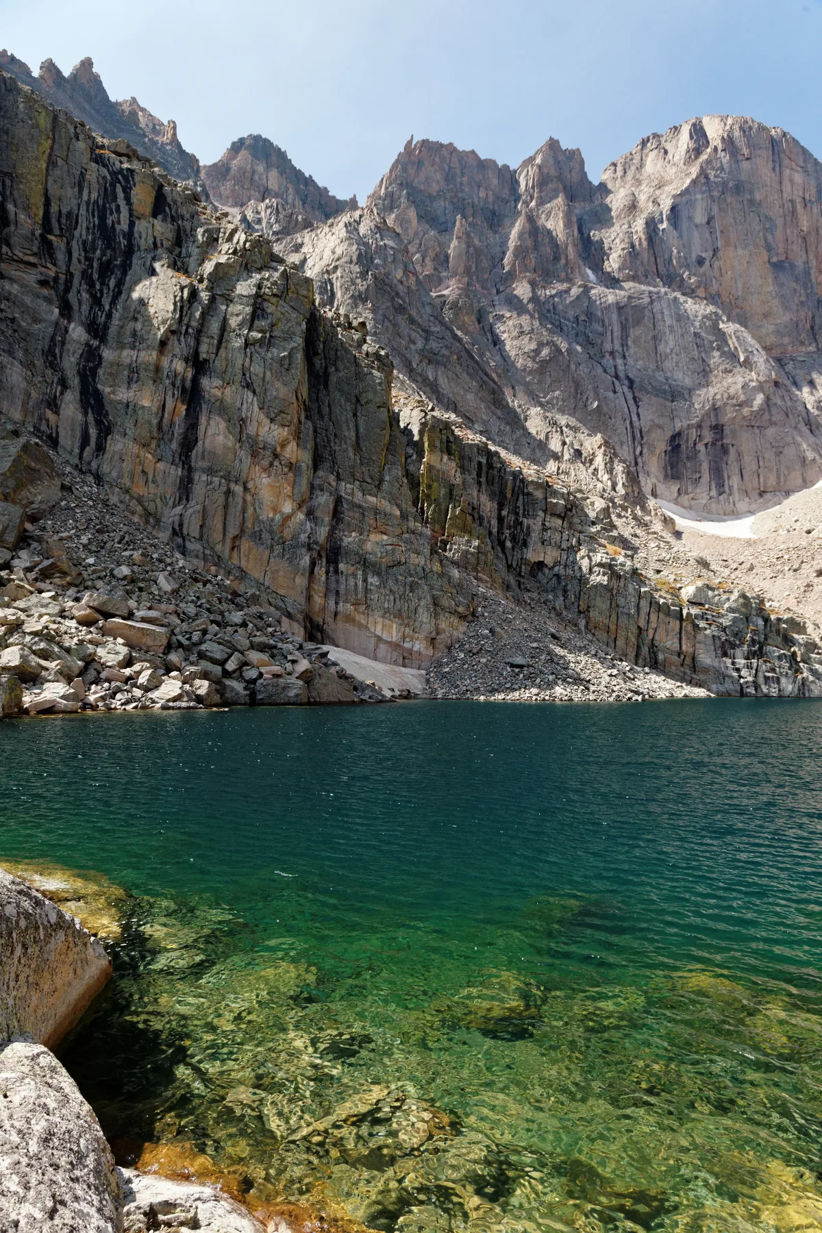 Clear emerald water fills the foreground of Chasm Lake, with rocky shoreline and bright sunlight revealing submerged stones near the edge. Above the lake, steep gray cliffs and the towering granite face of Longs Peak rise sharply into a pale blue morning sky.