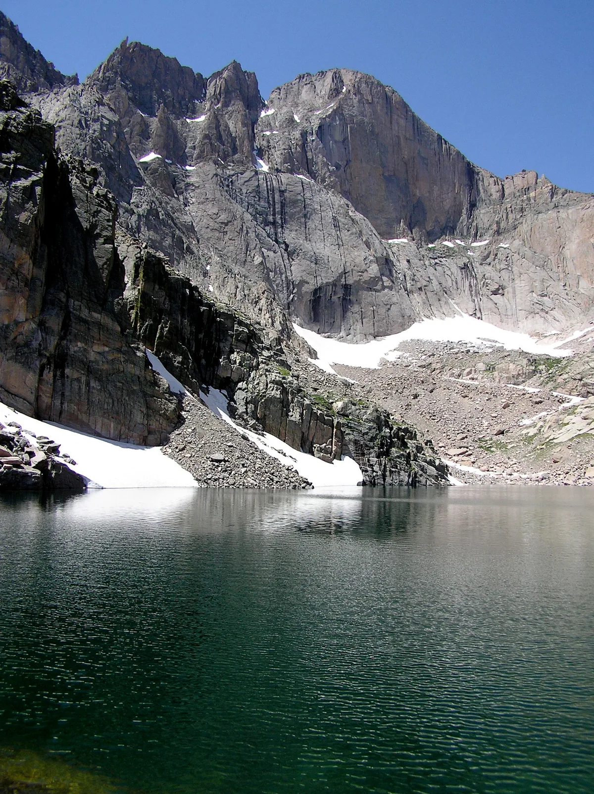 A calm alpine lake fills the foreground, its dark green water lightly rippled and reflecting the jagged granite walls above. Snow patches cling to the rocky cirque beneath Longs Peak’s sheer Diamond face under a clear blue morning sky.