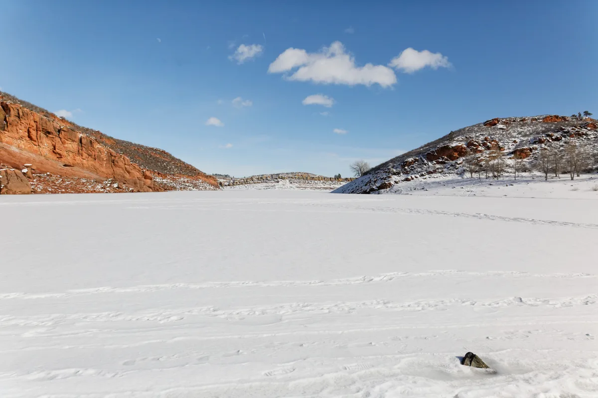 A frozen reservoir stretches across the foreground under a clear blue sky, with red rocky foothills and snow-dusted slopes rising on both sides. Thin tracks cross the ice, and a small rock pokes through the snow near the lower right edge.
