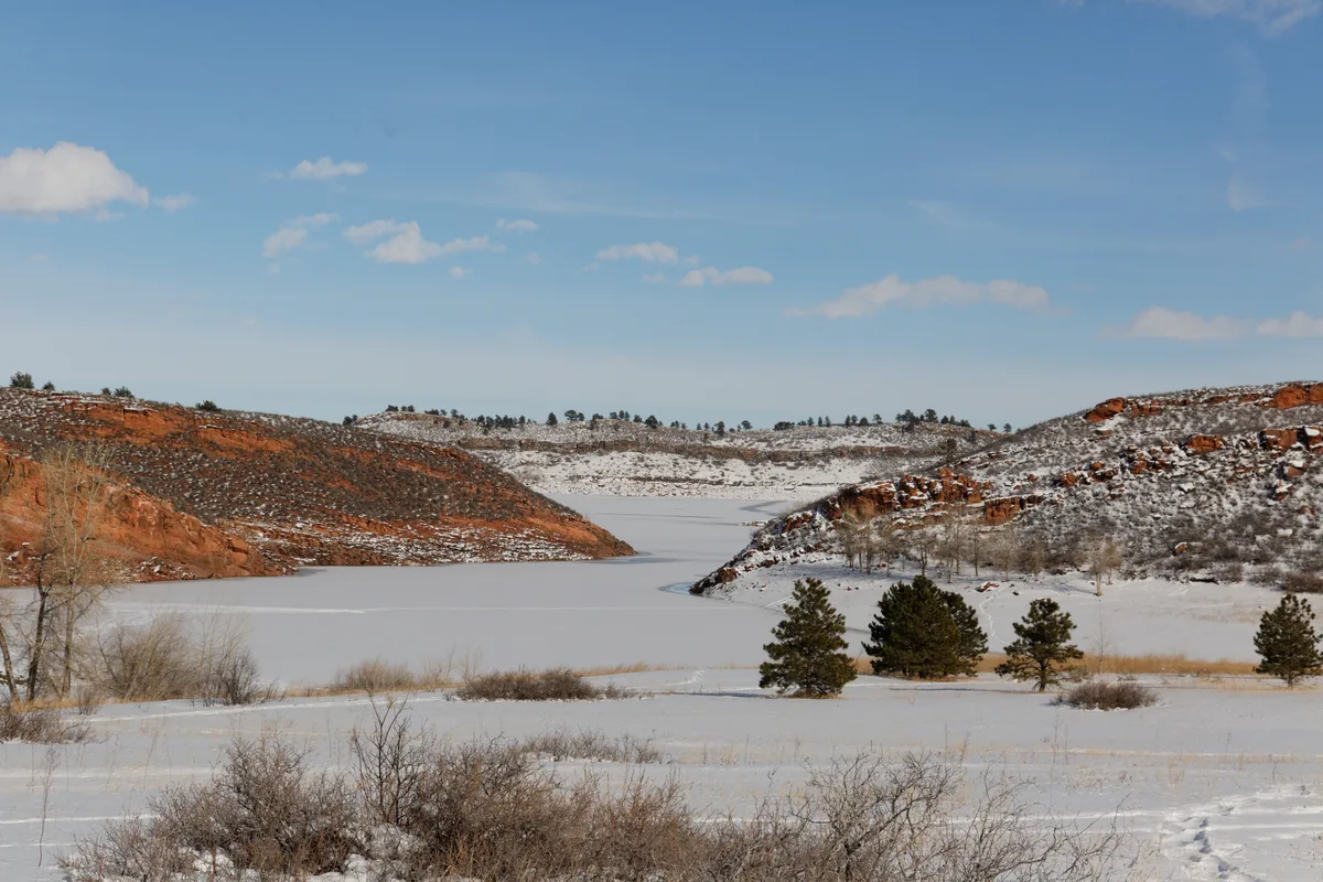 Snow blankets the frozen reservoir and open foreground at Lory State Park, with red sandstone hogbacks rising on both sides. Sparse pines and bare shrubs dot the white landscape beneath a clear blue sky with thin clouds.