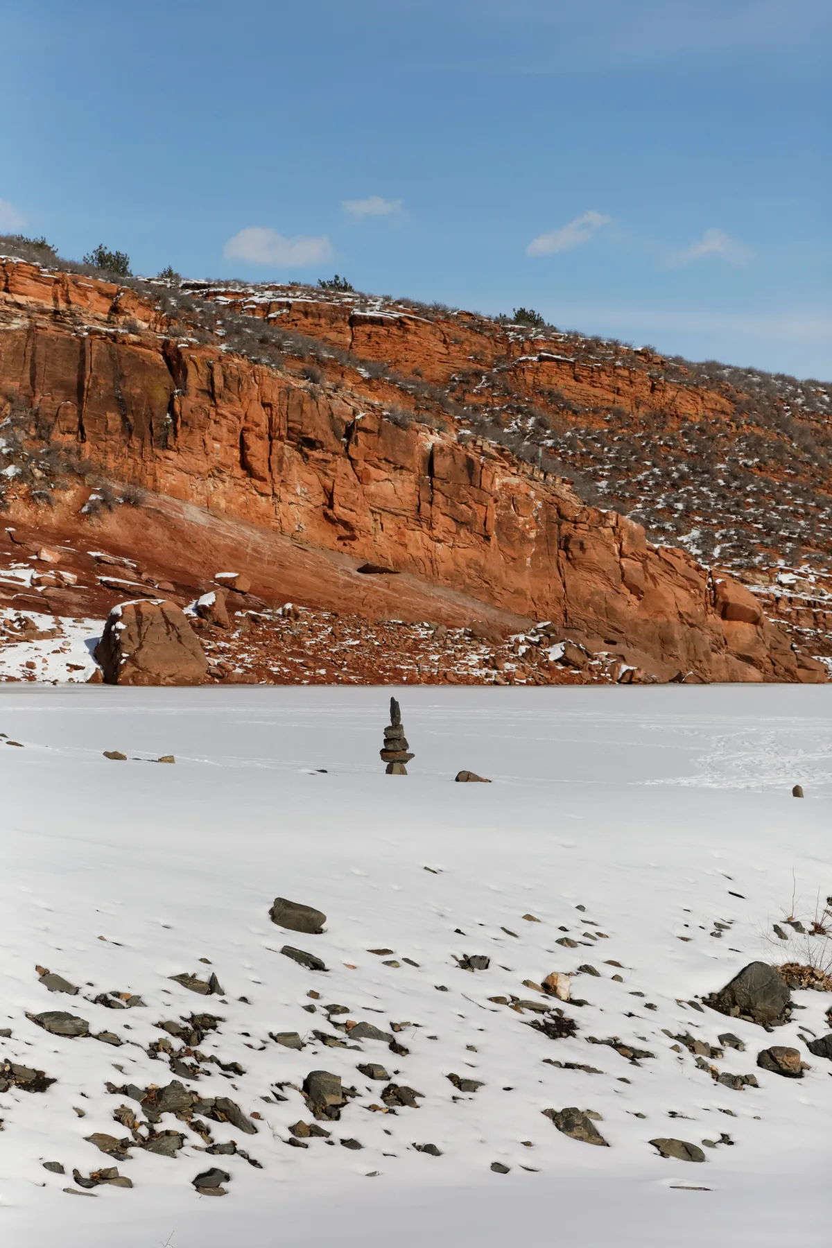 Red sandstone hogbacks rise behind a frozen, snow-covered foreground scattered with rocks, with a small stacked stone cairn near the center. Pale blue sky and thin clouds give the winter scene a clear, crisp feel.