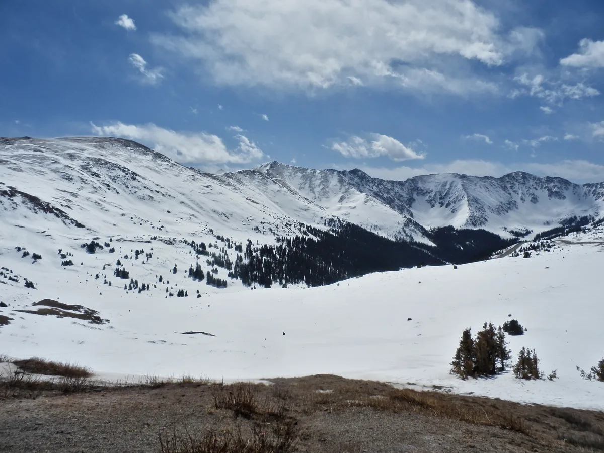 Snow-covered alpine slopes and a broad, white basin stretch below dark ridgelines at Loveland Pass, with scattered evergreens dotting the valley. A deep blue sky with thin clouds hangs over the high, windswept Continental Divide landscape.