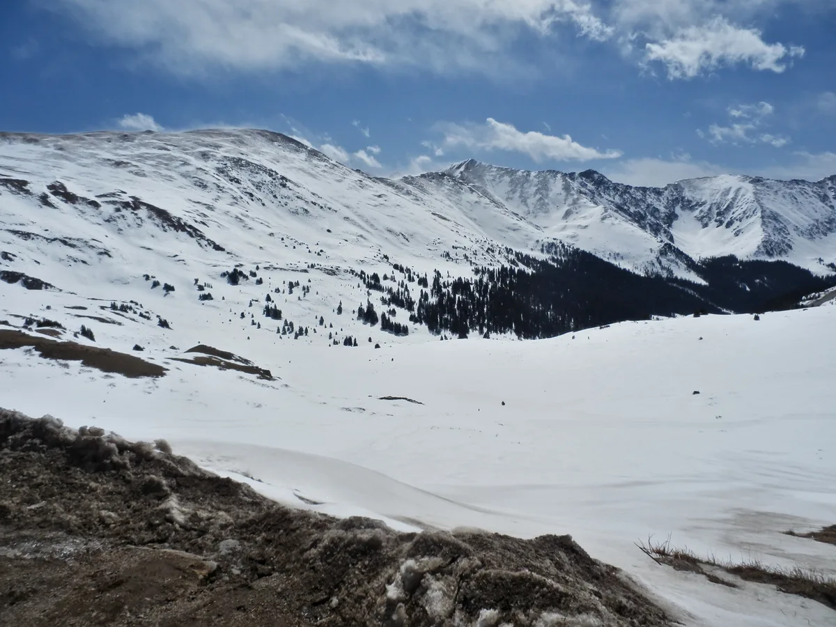 Snow-covered alpine slopes stretch across Loveland Pass beneath a blue sky with thin clouds, with dark bands of evergreens cutting through the valley and ridgeline. In the foreground, rough rocky tundra and wind-packed snow frame the wide mountain panorama.