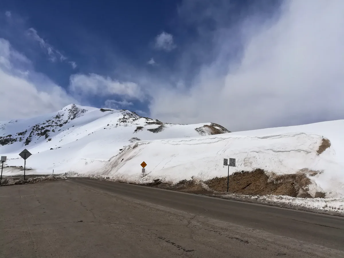 Snowy Loveland Pass curves past tall white drifts and exposed brown roadcuts, with rounded alpine ridges rising behind it. A deep blue sky with streaky clouds fills the top of the frame, emphasizing the high-elevation winter landscape.