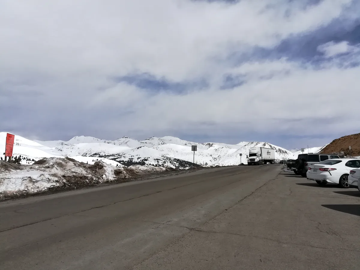 A mountain pass overlook with a paved road curving past parked cars and a white semi-truck, bordered by dirty spring snowbanks. Snow-covered alpine peaks stretch across the horizon beneath a bright, cloud-filled sky.