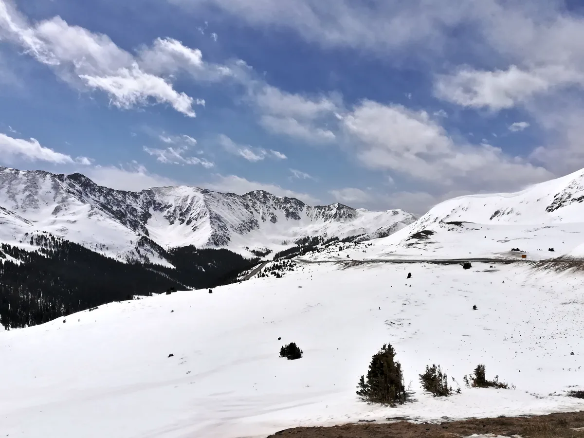 Snow-covered alpine tundra rolls down from Loveland Pass toward dark, rugged mountain ridges and a distant valley, with scattered evergreens breaking up the white slopes. Under a blue sky streaked with soft clouds, the high pass road cuts across the right side of the scene.