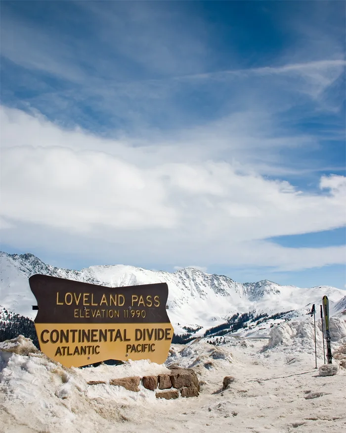 Loveland Pass sign stands in a snowy pullout at 11,990 feet, with the Continental Divide marker in the foreground and rugged white peaks stretching across the background. Thin clouds drift through a bright blue sky above the alpine ridgeline and snow-packed pass.