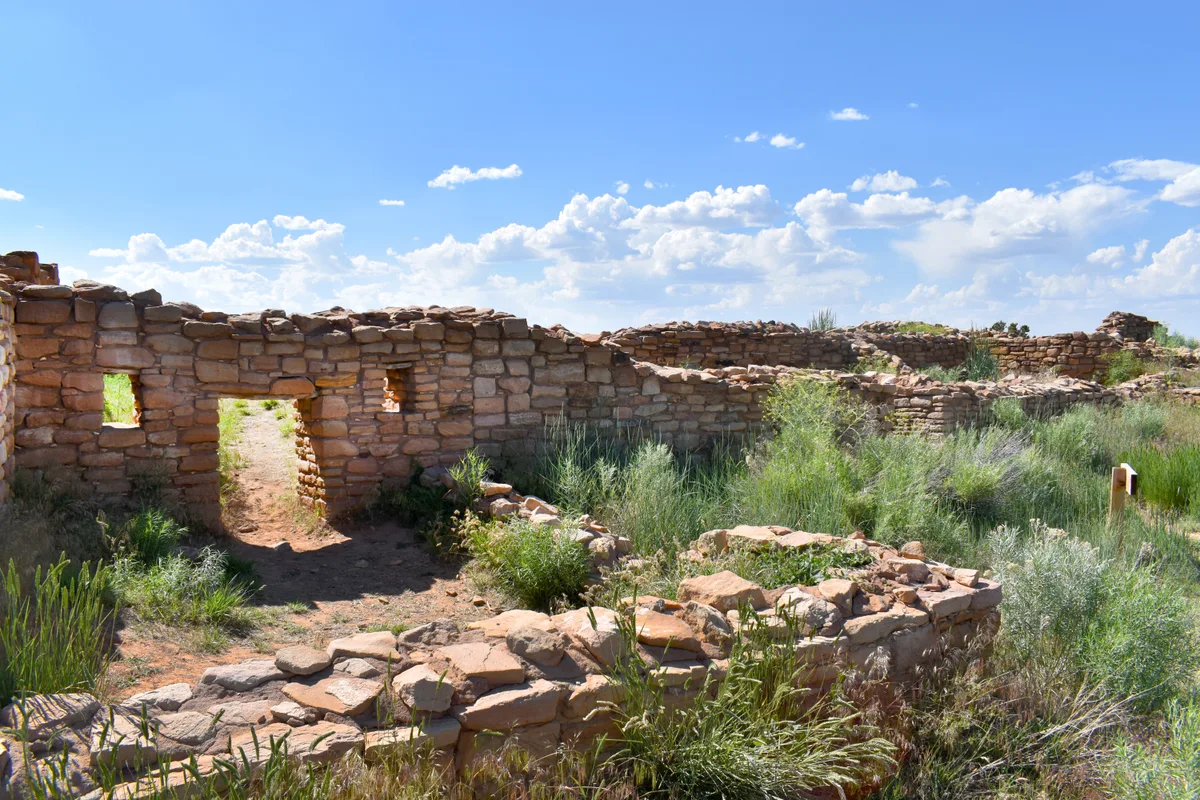 Sunlit stone ruins of Lowry Pueblo stretch across a sage-green plain, with low masonry walls and doorways framing a dirt path into the site. Bright blue sky and scattered white clouds open the scene, emphasizing the solitude of the ancient great house.