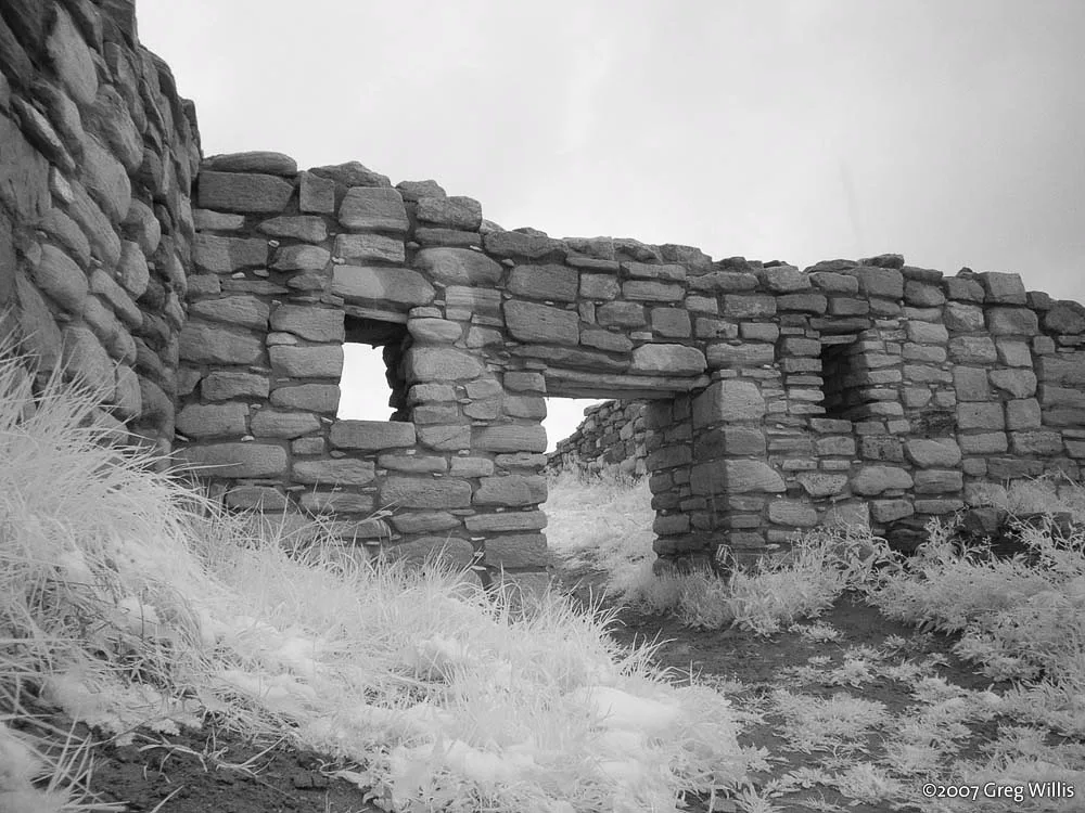 Stone Ancestral Puebloan walls frame a doorway and small window opening, with rough masonry blocks stacked against a bright sky. Pale sagebrush and grasses fill the foreground, giving the ruin a quiet, isolated feel.