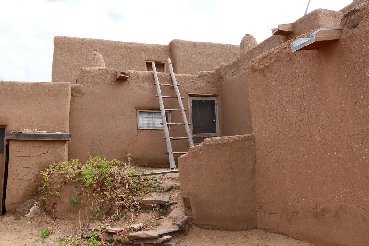 Adobe pueblo walls and stepped rooflines frame a small interior courtyard, with a weathered wooden ladder leaning up to a doorway and window. The sunlit earth-toned plaster shows hairline cracks and rough texture, with a few green plants growing beside the stone steps.