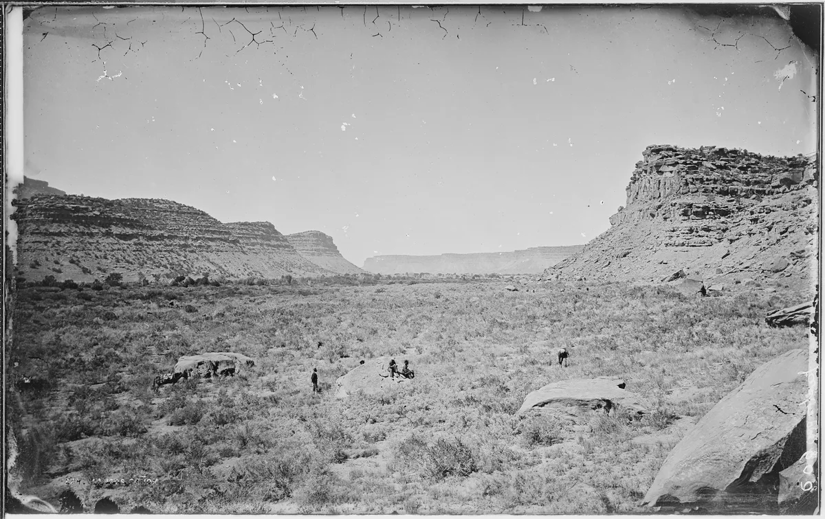 A wide black-and-white view of a broad desert canyon framed by layered mesa cliffs on both sides, with a few tiny figures and pack animals scattered across the grassy floor. The open sky and distant plateau create a deep, sunlit sense of scale and isolation.