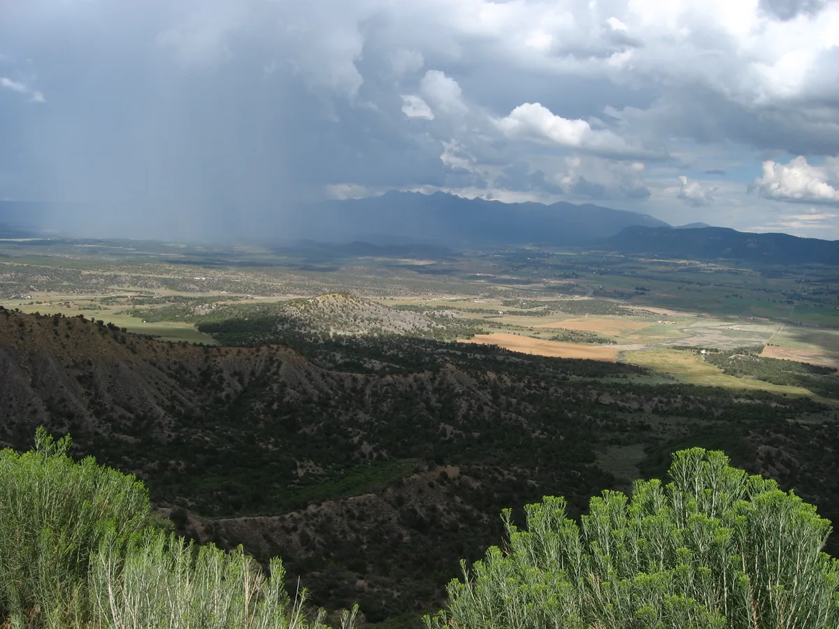 A wide overlook shows a storm sweeping across the valley, with a gray rain curtain dropping on the left and heavy clouds piling over distant mountains. Sunlight breaks through in patches to light the patchwork fields below, while green shrubs frame the foreground and dark ridges lead into the horizon.