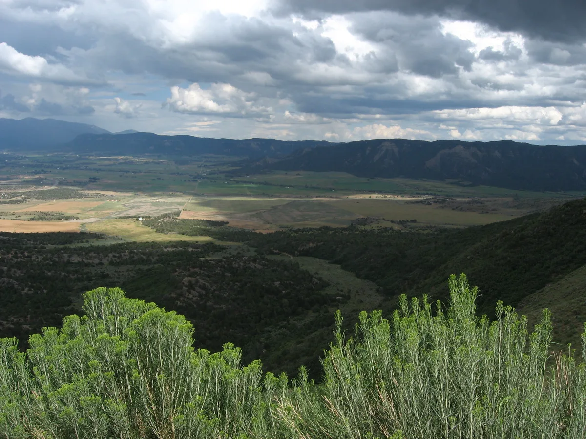 A wide overlook frames a broad green valley with patchwork fields, dark forested hills, and distant blue mountains under a sky filled with thick, dramatic clouds. Sunlight breaks through in spots, lighting the low shrubs in the foreground and parts of the valley below.