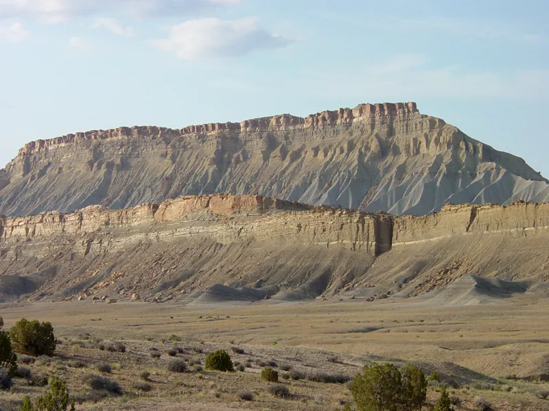 Layered shale cliffs rise above a broad, dry valley, with gray and tan slopes etched by erosion and capped by a flat, sunlit rim. Sparse shrubs and small trees dot the foreground under a pale blue sky with soft clouds.