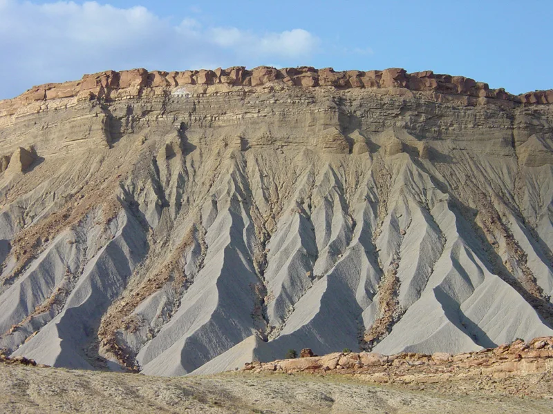 Steep gray shale badlands rise in sharp, eroded ridges below a flat, rust-colored cap of rock, with long shadows emphasizing the layered texture. The scene is lit by soft golden-hour light against a clear blue sky.