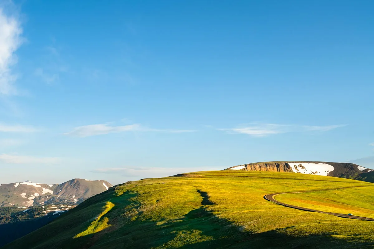 Golden hour light washes over a broad grassy ridge beside a curving mountain road, with dark green shadow bands stretching across the foreground and snow-patched peaks rising in the distance. The sky is clear blue with a few thin clouds, giving the overlook a wide, open feel.