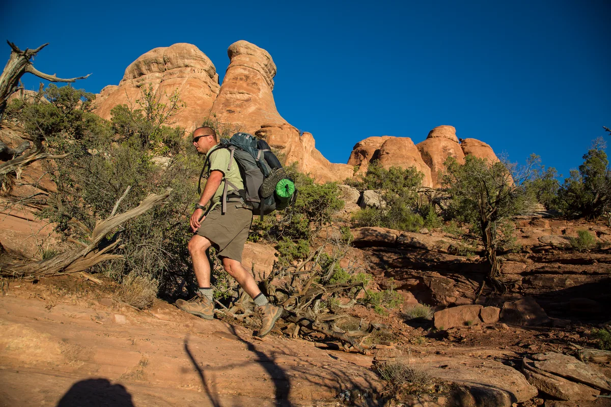 A hiker with a large backpack climbs a sunlit sandstone slope in Knowles Canyon, surrounded by scrub and deadwood under a deep blue sky. Towering orange Wingate rock formations rise behind him in the late-afternoon light.