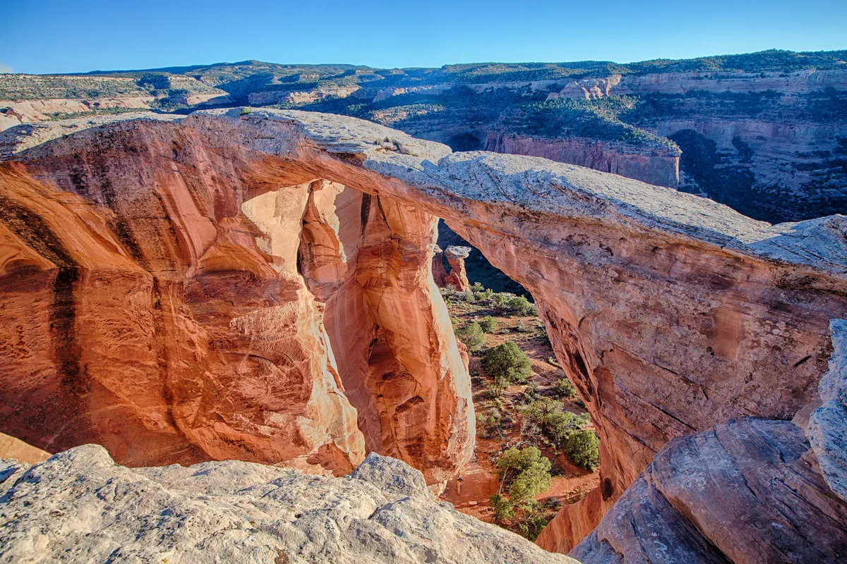 Sunlit Wingate sandstone forms a massive natural arch over a narrow desert canyon, with warm orange walls framing the shadowed drop below. The scene looks out across layered mesas and scrub-covered cliffs in clear afternoon light.