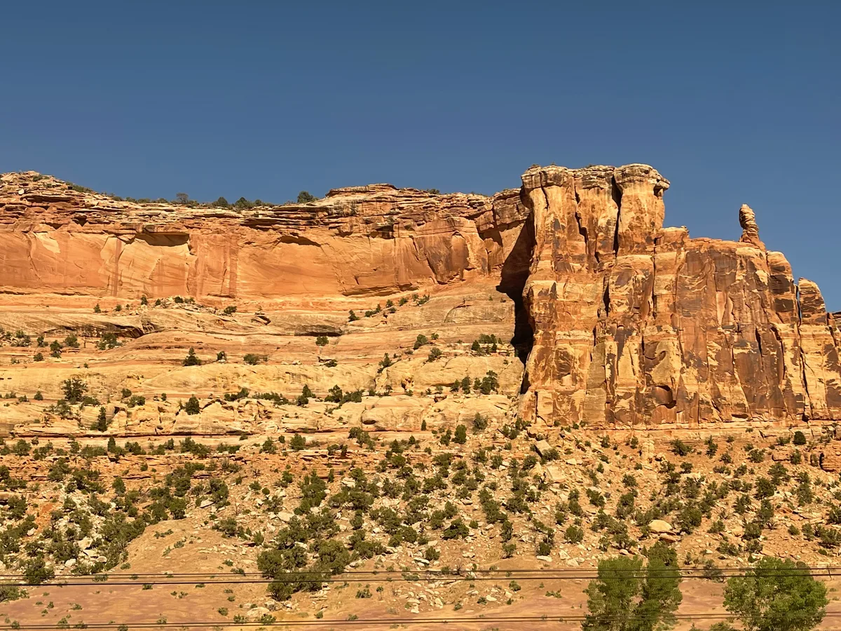 Sunlit orange sandstone cliffs rise above a sparsely vegetated desert slope, with a sheer rock tower and layered canyon walls set against a clear blue sky. The wide view is seen from a moving train, with a few dark utility lines crossing the bottom edge.