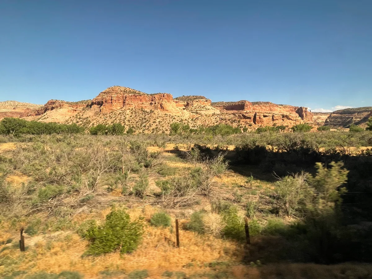 Sunlit red and tan sandstone cliffs rise above a scrubby desert foreground, with low green shrubs and dry grasses stretching across the valley. The scene is seen in wide daylight under a clear blue sky, with dark shadowed patches cutting across the lower right.