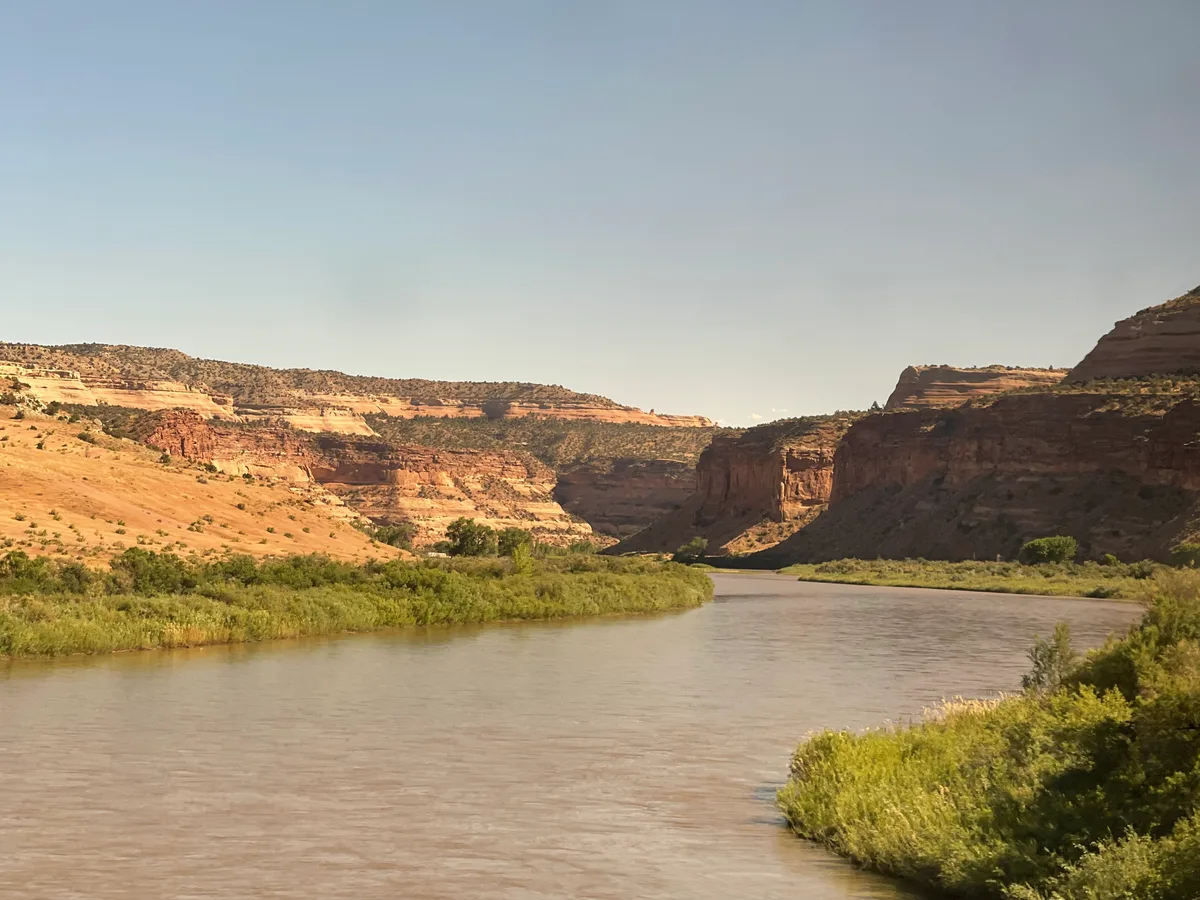 A broad river winds through the desert canyon, bordered by green cottonwood and brush, with sunlit layered sandstone cliffs rising on both sides. The warm afternoon light softens the red and tan rock under a clear blue sky.