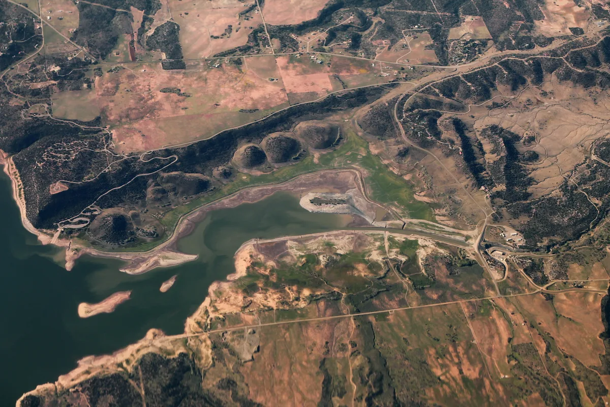 An aerial view of McPhee Reservoir shows dark green water edged by pale, muddy shorelines and small exposed islands, with winding roads and patchwork fields spreading across the surrounding mesa. The late-day light warms the tan and rust-colored terrain, emphasizing the reservoir’s broad, rugged desert setting.