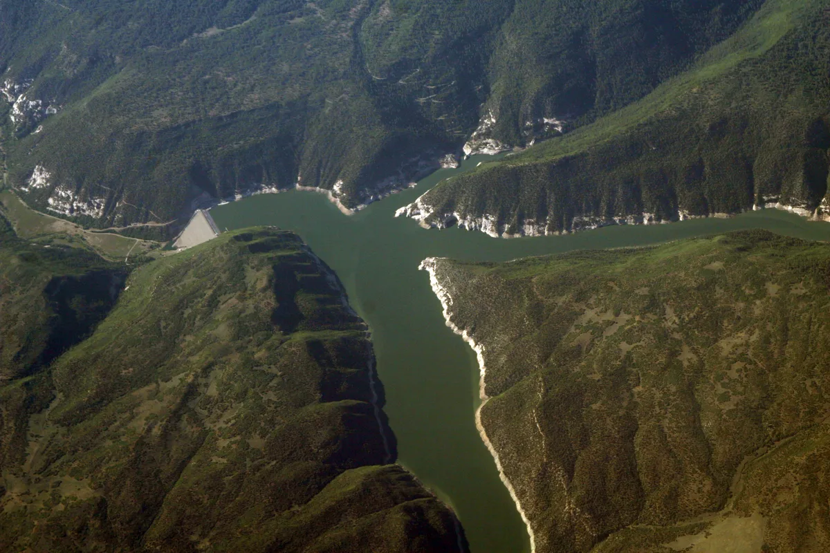 A wide aerial view shows McPhee Reservoir winding through steep, green-brown canyon walls, with pale rocky shoreline edges tracing the water. Warm golden light and soft shadows emphasize the rugged mesa terrain and the reservoir’s forked, reflective channels.