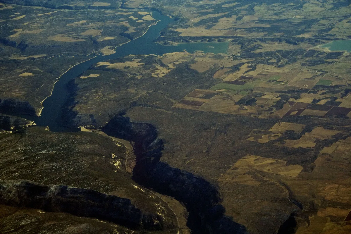 A high aerial view shows McPhee Reservoir stretching through a broad, rugged landscape, with dark blue water cutting between shadowed cliffs and patchwork fields below. The golden light picks out the shoreline and dry mesa terrain, creating strong contrast between the reservoir and the surrounding desert land.