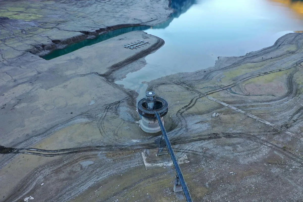 An aerial view shows McPhee Reservoir at very low water, with a circular intake tower and long blue access bridge standing on exposed, muddy shoreline. The still reservoir surface reflects pale sky light, while dark tire tracks and layered sediment patterns curve across the dry lakebed.