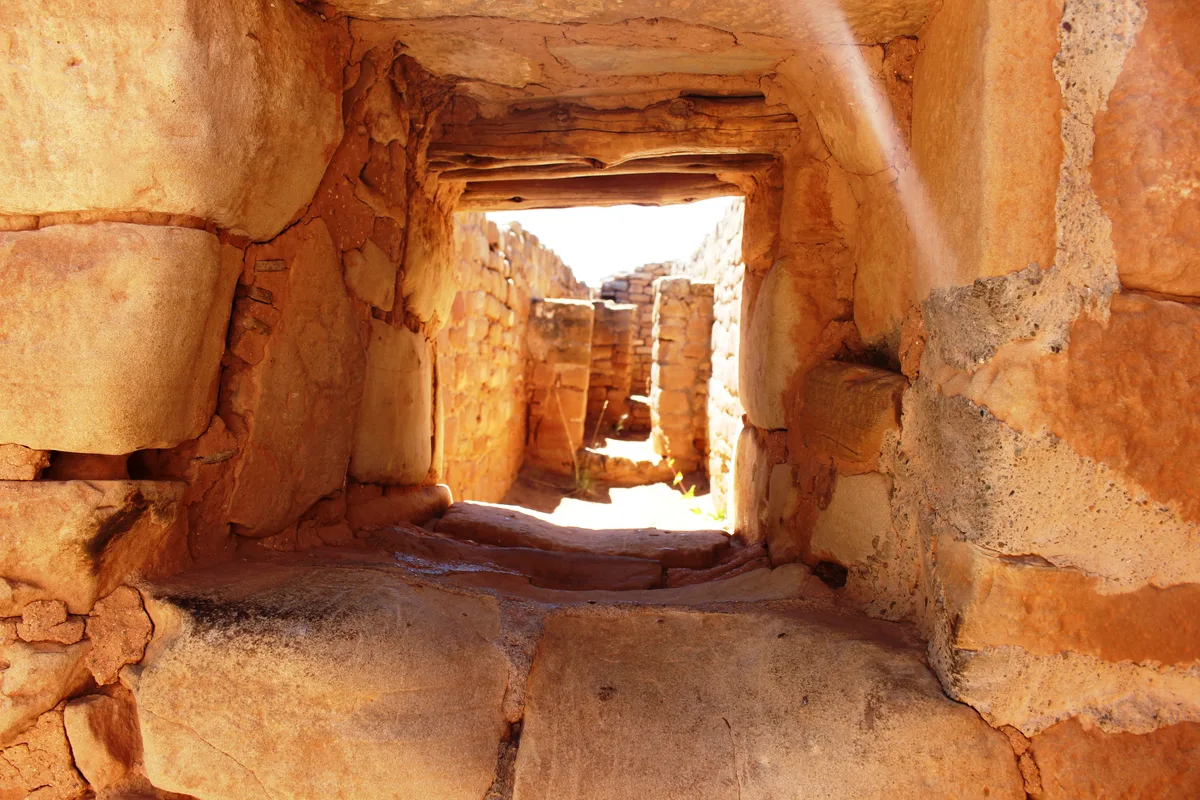 Sunlit sandstone masonry forms a narrow passage or doorway, with rough stacked stone walls and a timber lintel framing a bright opening beyond. Warm afternoon light brings out the orange and tan textures of the ancient construction.