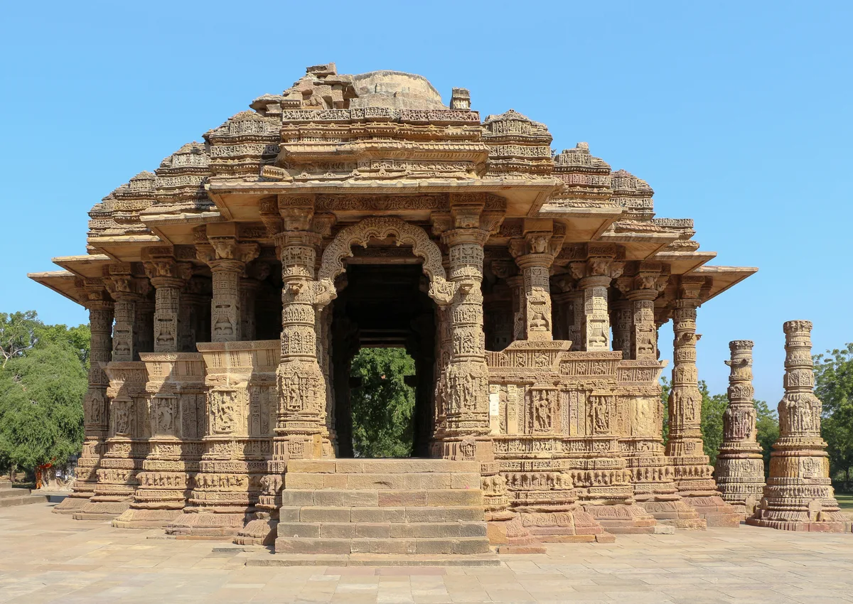 Sunlit sandstone Sun Temple with richly carved pillars, layered rooflines, and a wide central doorway, set on a paved terrace under a clear blue sky. The afternoon light brings out the detailed masonry and the freestanding columns at the right edge.