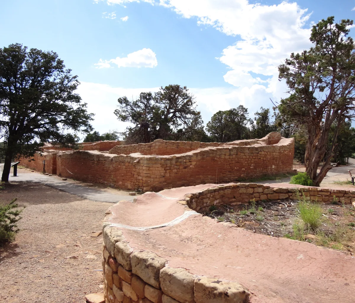 Weathered red-and-tan stone walls of Mesa Verde’s Sun Temple curve through the foreground and middle ground, with the unfinished D-shaped masonry enclosure surrounded by juniper trees. Bright afternoon light and a blue sky with scattered clouds emphasize the texture of the ancient stonework and the open mesa-top setting.